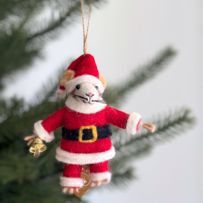 A playful felt Santa Mouse ornament hangs from a Christmas tree branch by a jute string. This handcrafted wool felt ornament features a white-faced mouse with beige ears and long black whiskers. Dressed in a classic red Santa suit, trimmed with white faux fur and cinched by a black belt with a gold buckle, the Santa mouse holds a tiny gold bell.
