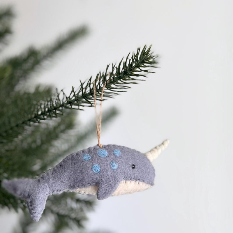 A felt narwhal Christmas ornament hangs gracefully from a green Christmas tree branch. This unique handcrafted felt ornament features a gray body adorned with light blue polka dots, a white underbelly, and a distinct white horn, suspended by a natural jute string.