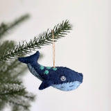 A handcrafted felt whale ornament, from the Felt Sea Animal Ornaments set, hangs charmingly from a green Christmas tree branch against a light background. This nature-inspired felt whale ornament is navy blue on its upper body with a lighter blue underside, detailed with green spots, a fin, tail, and a visible eye. It is suspended by a jute string.