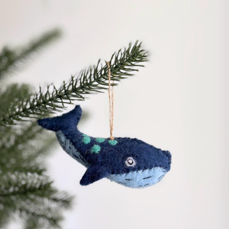 A handcrafted felt whale ornament, from the Felt Sea Animal Ornaments set, hangs charmingly from a green Christmas tree branch against a light background. This nature-inspired felt whale ornament is navy blue on its upper body with a lighter blue underside, detailed with green spots, a fin, tail, and a visible eye. It is suspended by a jute string.