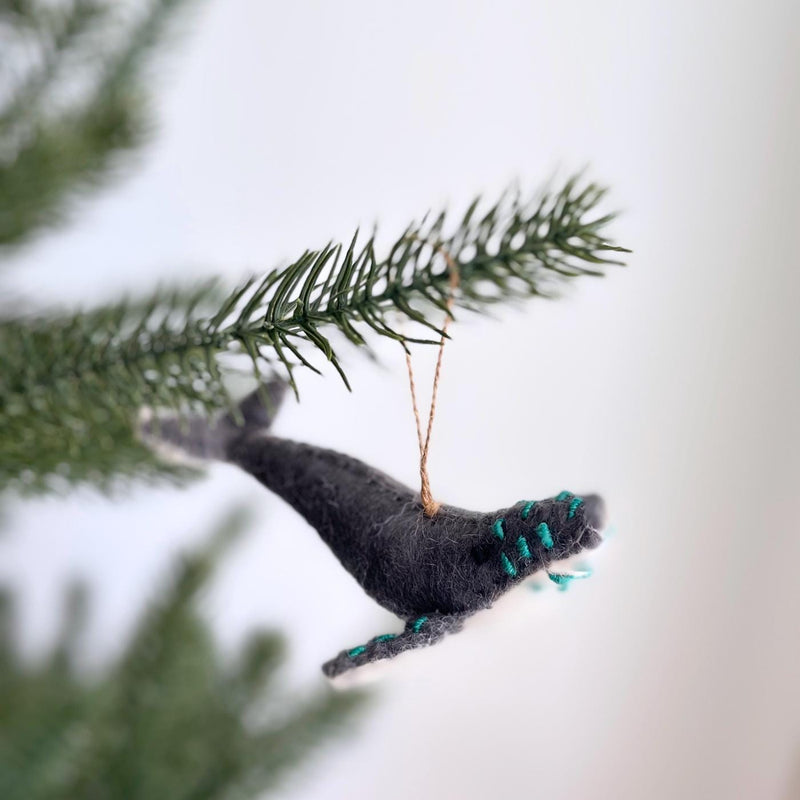 A gray wool felt whale ornament, part of a set of felt sea animal ornaments, hangs on a Christmas tree. This handmade felt whale ornament features a gray body with a turquoise-embroidered tail. A visible jute string on its back allows for hanging, showcasing its detailed texture and craftsmanship.