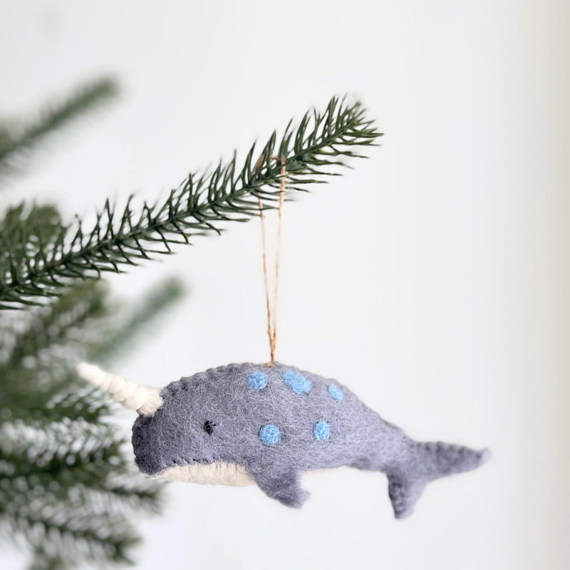 A gray felt narwhal ornament hangs from a lush green pine branch against a soft gray backdrop. This handmade felt narwhal features a white horn and belly, light blue felt circles, and a small black eye. Its textured material gives it a cozy feel, suspended by a jute string, adding a unique, handcrafted touch to holiday decor.