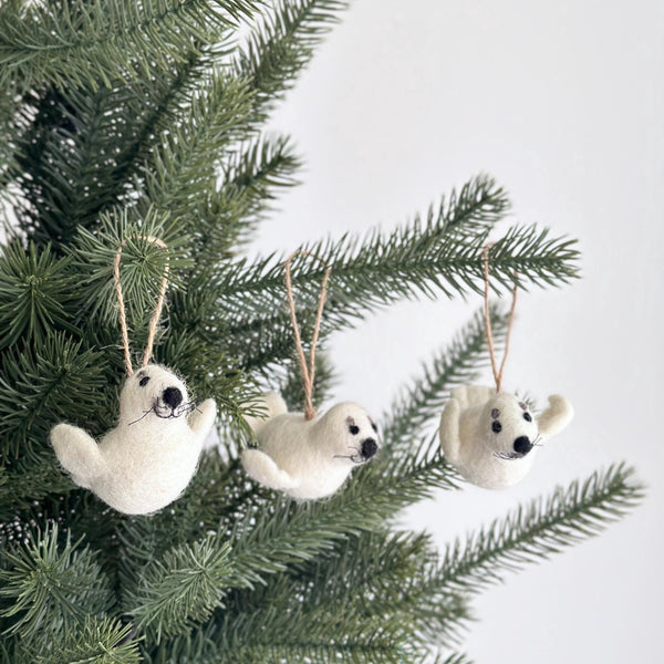 Three white wool felt seal ornaments are playfully displayed on a Christmas pine tree, each suspended by a natural hemp string. These handcrafted felt seal ornaments feature distinct black eyes and a charming stitched nose, adding a festive touch.