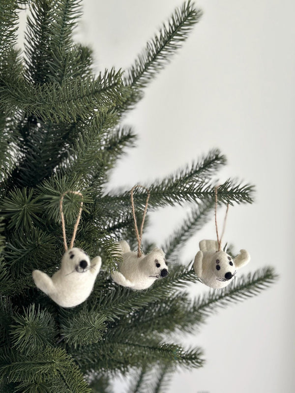 A set of three white needle-felted wool felt seal ornaments hang from thin hemp strings on a close-up section of a Christmas pine tree branch. Each handcrafted seal features black eyes, whiskers, slightly raised flippers, and a stitched smile, embodying ethical craftsmanship.