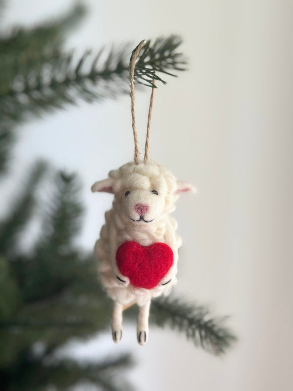 A wool felt sheep holding heart ornament rests on a blurred green pine branch. This soft, off-white ornament features a rounded, tufted body, floppy ears, black eyes, a pink nose, and a stitched mouth, creating a gentle expression. The sheep holds a small red heart, and a jute string is attached for easy hanging.