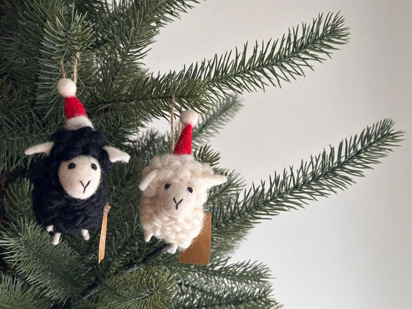 Two cozy wool felt sheep ornaments with Christmas hats, one black and one white, are nestled among vibrant green pine needles. Each ornament features fuzzy wool fur, a cream-colored face with a stitched 'Y' mouth, and a small red and white Santa hat. The white sheep also wears a colorful scarf, both ready to hang with a hemp string loop, evoking a festive mood.