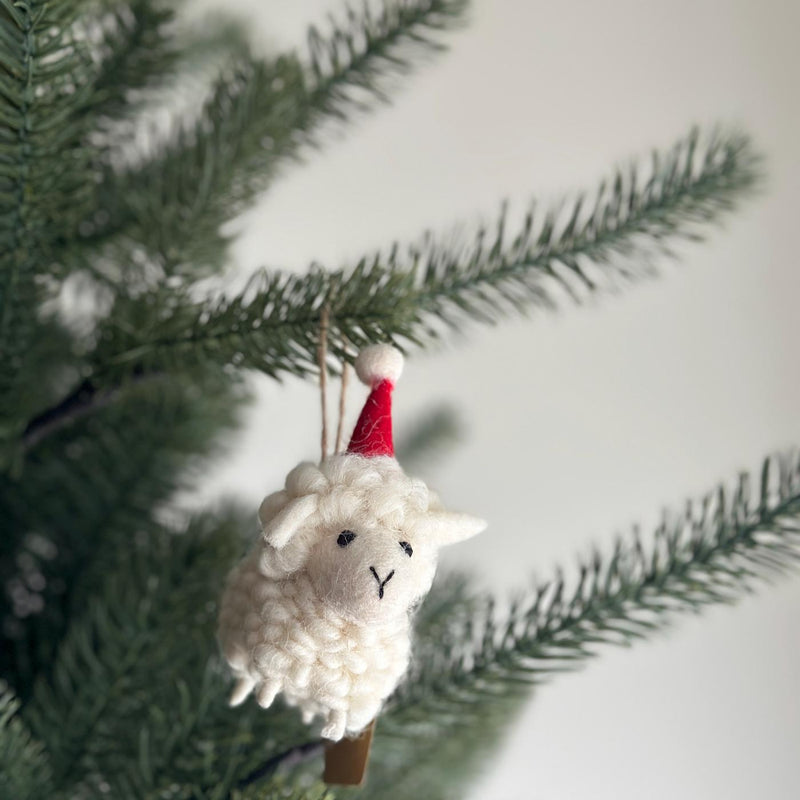 A wool felt sheep ornament with a Christmas hat hangs on a Christmas tree. This charming handmade felt ornament features a soft, fluffy off-white body, small black eyes, and a stitched black 'Y' mouth. A tiny, pointed red Christmas hat with a white pompom adorns its head, and a hemp string loop is attached for easy hanging.