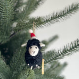 A fuzzy black and white felt sheep ornament with a red and white Christmas hat hangs on a dark green Christmas tree branch. The handcrafted wool felt ornament has tiny white feet and a simple face with a stitched 'Y' mouth, ready to adorn your tree.