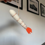 A hand holds an adorable, handcrafted felt shrimp ornament, suspended by a jute string, against a white wall with framed pictures. This felt shrimp ornament is off-white with light pink bands around its body and a vibrant orange tail, and it contains bendable wire for interactive posing.