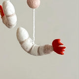 A whimsical white wool felt shrimp ornament, featuring subtle gray stripes and a vibrant red tail, floats suspended in mid-air. Attached to this delicate felt shrimp ornament is a white string leading upwards to a fuzzy light brown felt ball. This playful handcrafted felt ornament adds a charming, sea-themed touch.