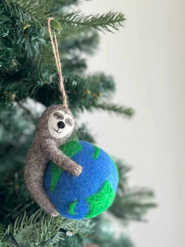 A gray wool felt sloth ornament hangs from a green Christmas tree branch, suspended by a jute string. The wool felt sloth, featuring closed eyes and a stitched smile, gently embraces a blue and green globe showing landmasses on an ocean. This unique felt sloth Christmas ornament is a delightful decoration.