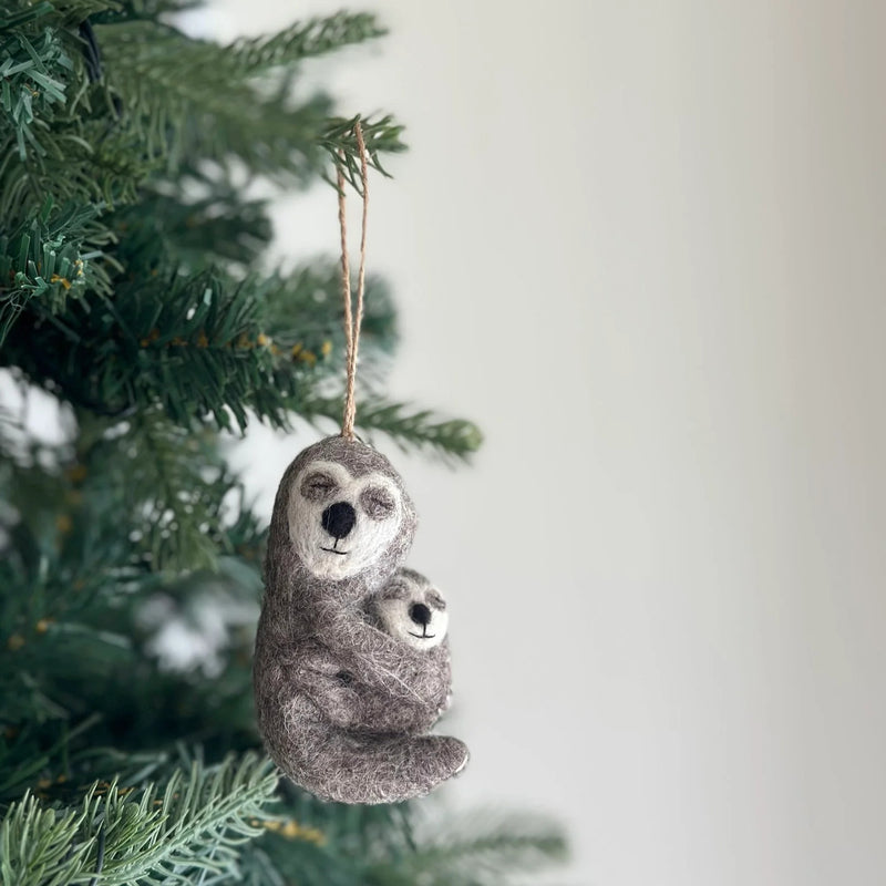 A wool felt sloth ornament, depicting a gray mother sloth with a white face, black nose, and closed eyes tenderly embracing her smaller baby, hangs from a green Christmas tree branch. This handcrafted felt Christmas tree ornament, made from textured gray wool felt, features a jute string for hanging and evokes a sense of warmth and familial love.