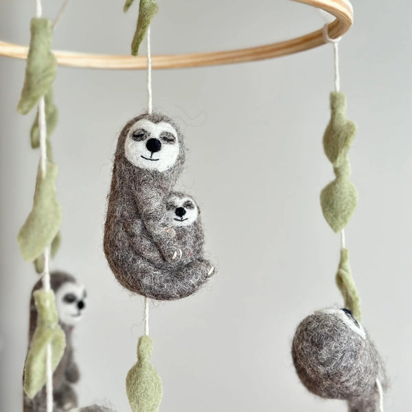 Felt Sloth Hugging Baby ornaments are displayed hanging from a wooden mobile frame. The prominent gray and cream felt sloth ornament features a serene expression with closed eyes and a gentle smile, tenderly cradling a smaller baby sloth, also with closed eyes. White cotton strings run through light green felt leaf embellishments, attaching the ornament to the frame. Two additional felt sloth ornaments are partially visible in the soft, light background.