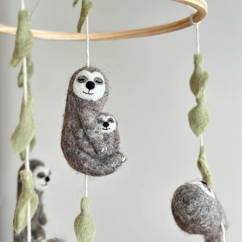Felt Sloth Hugging Baby ornaments are displayed hanging from a wooden mobile frame. The prominent gray and cream felt sloth ornament features a serene expression with closed eyes and a gentle smile, tenderly cradling a smaller baby sloth, also with closed eyes. White cotton strings run through light green felt leaf embellishments, attaching the ornament to the frame. Two additional felt sloth ornaments are partially visible in the soft, light background.