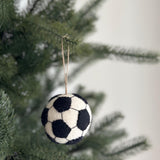 A handmade wool felt soccer ball ornament hangs from a Christmas tree branch, set against a soft, blurred festive backdrop. The round ornament is white with black pentagons stitched across its surface, mimicking a classic soccer ball design. A jute string is attached to the top, allowing it to dangle naturally among the lush green needles.