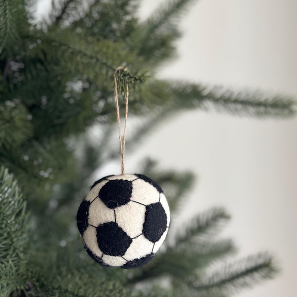 A handmade wool felt soccer ball ornament hangs from a Christmas tree branch, set against a soft, blurred festive backdrop. The round ornament is white with black pentagons stitched across its surface, mimicking a classic soccer ball design. A jute string is attached to the top, allowing it to dangle naturally among the lush green needles.