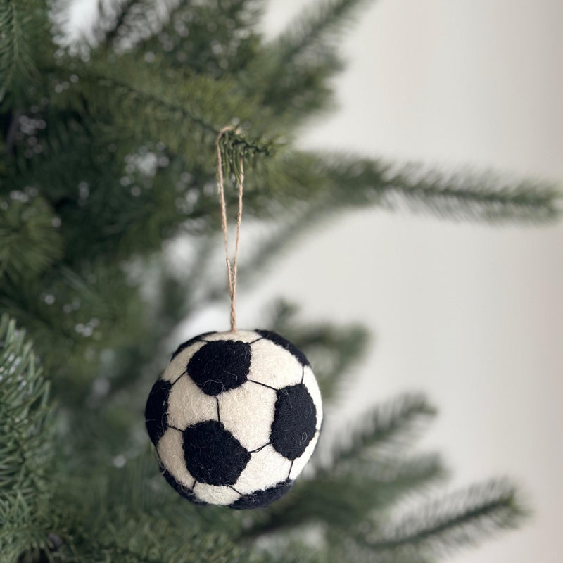A handmade wool felt soccer ball ornament hangs from a Christmas tree branch, set against a soft, blurred festive backdrop. The round ornament is white with black pentagons stitched across its surface, mimicking a classic soccer ball design. A jute string is attached to the top, allowing it to dangle naturally among the lush green needles.
