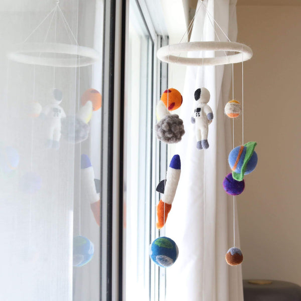 A colorful wool felt spaceman baby mobile hangs suspended in front of a bright window with white curtains, creating a soft, dreamy atmosphere. The mobile features a white felt ring from which a white spaceman, colorful planets, an orange and blue rocket, and a gray asteroid are suspended. Each needle-felted figure, crafted from 100% New Zealand wool, is detailed and textured, offering a whimsical, three-dimensional look.