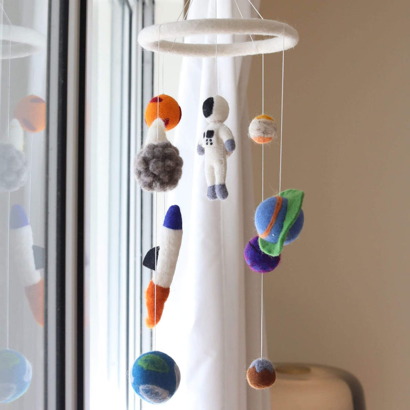A felt spaceman baby mobile is displayed in a softly lit room by a window with white curtains. This baby crib mobile features a white felt ring suspending by clear strings a white spaceman with black and gray details, a gray rocket with blue and orange accents, and colorful planets. The planets vary in size and texture, in orange, blue, green, and purple, evoking a sense of wonder for a nursery.