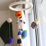 A wool felt spaceman baby mobile hangs brightly lit in front of a window, suggesting an airy nursery. It features a circular felt frame from which celestial-themed, needle-felted 3D figures are suspended. These include a white astronaut, colorful planets, a comet with a grey and yellow tail, and a white and orange rocket with "USA" lettering.