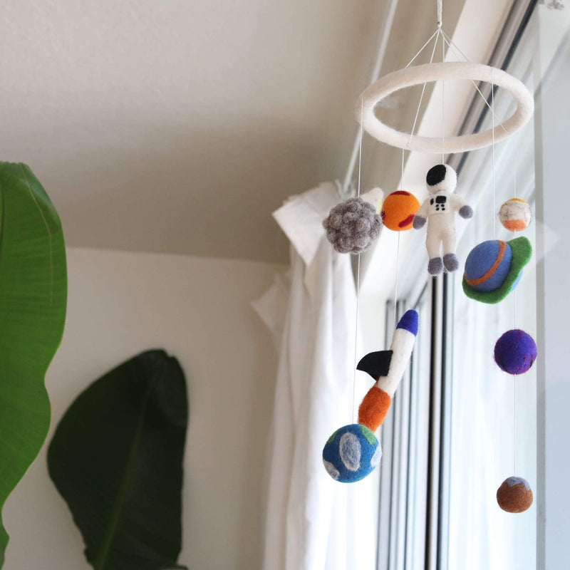A wool felt spaceman baby mobile hangs in a bright, airy room next to a window, against white walls and a sheer white curtain. The mobile features several 3D needle-felted figures, including a white astronaut, a blue and green rocket, and planets in shades of grey, orange, blue, green, purple, and brown, suspended from a white felt ring. A large green tropical leaf in the foreground adds a natural touch, evoking a sense of wonder.