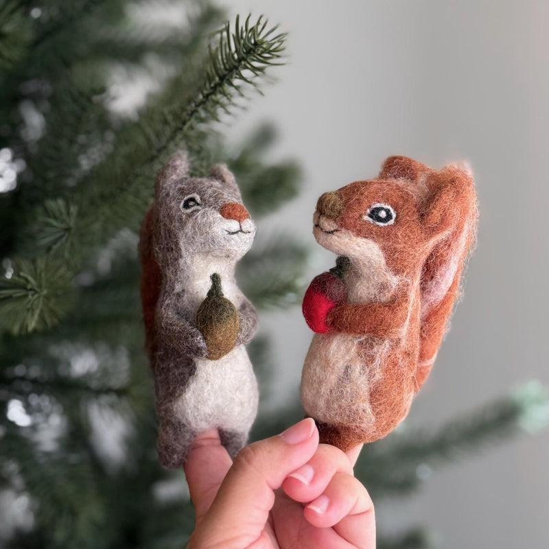 Two handcrafted needle felted squirrel finger puppets, known for their eco-friendly design, are playfully held by a female hand against a blurred green pine tree. These unique felt squirrel finger puppets, made by artisans, feature one gray squirrel holding a tiny brown acorn and another reddish-brown squirrel clutching a bright red nut. Both showcase small black eyes and intricate textures, perfect for imaginative woodland storytelling.