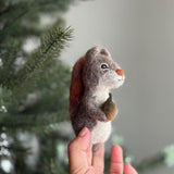 A gray, brown, and white handmade felt squirrel finger puppet is gently held in a female hand. Crafted from soft wool felt, this adorable squirrel puppet features a small, smiling face and clutches a tiny brown acorn with a green top, set against a blurry, festive background with a Christmas tree.