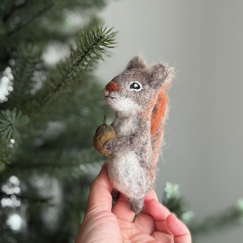A handmade felt squirrel finger puppet, crafted from wool felt, is held in a female hand against a softly blurred Christmas pine tree background. The gray squirrel puppet features a reddish-brown bushy tail and clutches a small brown acorn in its paws. This charming, needle-felted toy highlights intricate artisan detail, offering an eco-friendly option for imaginative play.