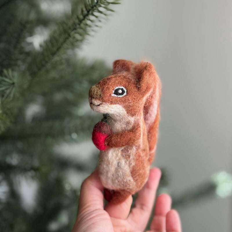 A handcrafted felt squirrel finger puppet, holding a tiny red acorn, is presented in a female hand against a blurred Christmas tree background. This unique wool felt finger puppet is meticulously needle-felted by artisans, showcasing textured brown, white, and red fur, a bushy tail, small ears, and expressive eyes. It's perfect for imaginative woodland play and seasonal storytelling.