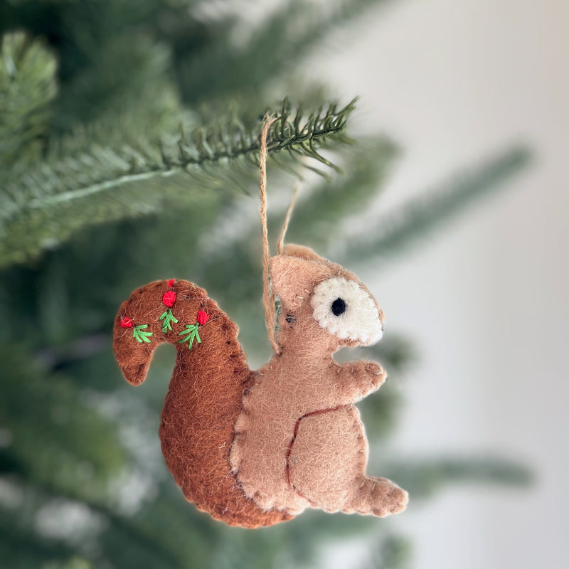 A wool felt squirrel ornament hangs on a Christmas tree, with blurred green pine needles in the background, creating a cozy holiday scene. This charming squirrel features a light brown body, a white face, and a bushy dark brown tail adorned with hand-embroidered red berries and green sprigs. A twine loop is attached for easy hanging, completing its rustic appeal.