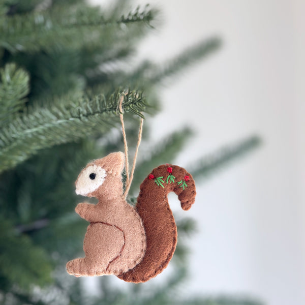 A light brown wool felt squirrel ornament hangs from a green Christmas tree branch. Its darker bushy tail is adorned with delicate hand embroidery featuring red berries and green sprigs. This handcrafted felt ornament adds a cozy, rustic woodland touch to holiday decor.