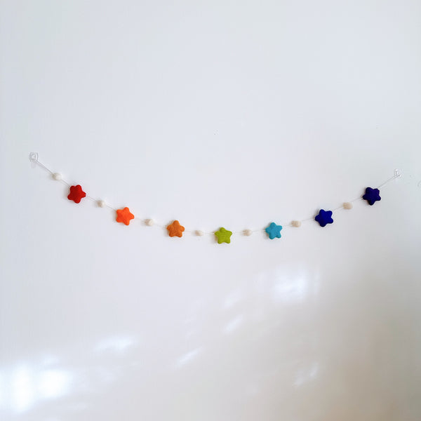 A felt star garland is displayed against a stark white background, creating a cheerful atmosphere. The garland features seven vibrant felt stars, each a different rainbow color: red, orange, yellow, green, light blue, dark blue, and navy blue.