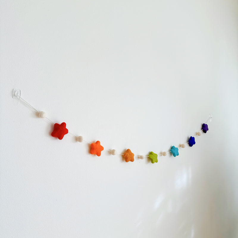 The colorful felt star garland is displayed on a stark white wall. This handmade felt garland features seven vibrant rainbow-hued felt stars, each separated by a small felt ball.