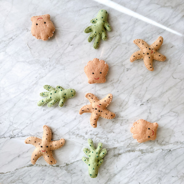 The wool felt Starfish Garland, displaying a whimsical coastal theme, rests on a white marble surface. It features alternating wool felt starfish, seaweed, and seashells connected by a thin white string. The starfish are light brown with dark brown spots, the seaweed is green with darker green spots, and the seashells are light brown with white accents and delicate stitching.