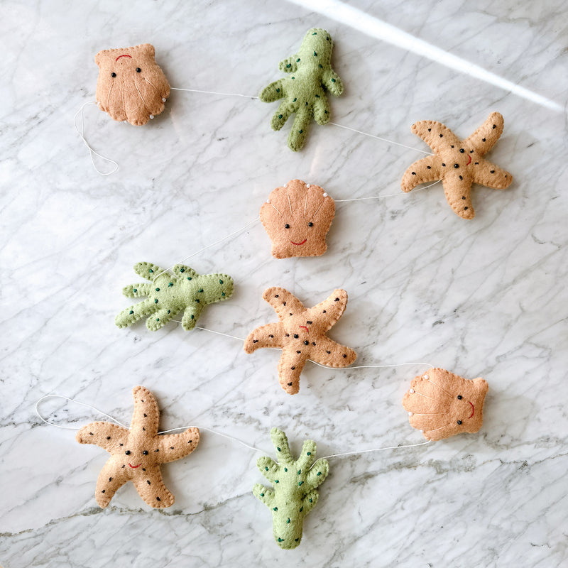 The wool felt Starfish Garland, displaying a whimsical coastal theme, rests on a white marble surface. It features alternating wool felt starfish, seaweed, and seashells connected by a thin white string. The starfish are light brown with dark brown spots, the seaweed is green with darker green spots, and the seashells are light brown with white accents and delicate stitching.