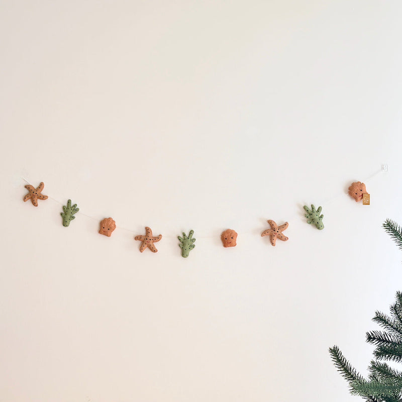 A wool felt starfish garland is displayed against a neutral wall, evoking a calm, coastal atmosphere. The garland features alternating tan felt starfish with dark brown stitching, green felt seaweed with dark green dots, and tan felt seashells with smiling faces. It is strung on a clear string and attached to the wall with a clear hook.