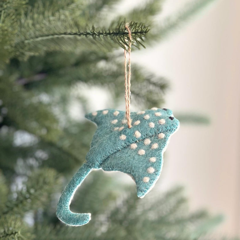 A handmade wool felt stingray ornament with a hemp string hangs on a festive green pine tree, creating a cheerful, artisanal atmosphere. The light teal stingray, featuring white polka dots, has a long curved tail and small black eyes, embodying an eco-friendly holiday spirit.