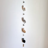 A charming wool felt cat garland, featuring stitched details, is displayed against a plain white wall. Four 3D needle-felted cats are strung vertically: a spotted white cat, a gray striped cat, a tan striped cat, and a solid black cat. A small copper bell hangs from the very bottom, completing the delightful design.