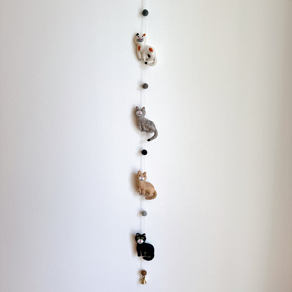 A charming wool felt cat garland, featuring stitched details, is displayed against a plain white wall. Four 3D needle-felted cats are strung vertically: a spotted white cat, a gray striped cat, a tan striped cat, and a solid black cat. A small copper bell hangs from the very bottom, completing the delightful design.