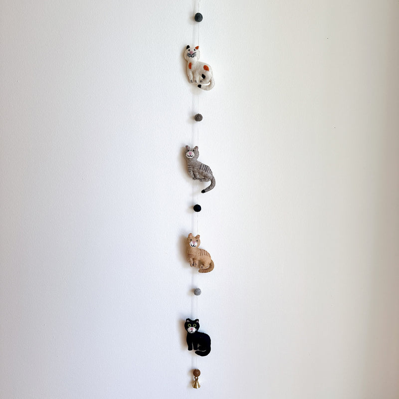 A charming wool felt cat garland, featuring stitched details, is displayed against a plain white wall. Four 3D needle-felted cats are strung vertically: a spotted white cat, a gray striped cat, a tan striped cat, and a solid black cat. A small copper bell hangs from the very bottom, completing the delightful design.