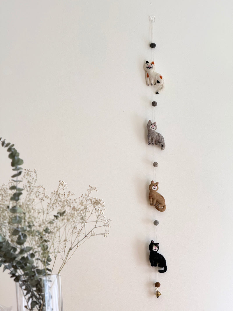 A charming felt cat garland hangs against a plain white wall, with a vase of baby's breath flowers partially visible to the lower left. The garland features four distinctive felt cats: one white with orange and black spots, one gray, one brown, and one black. Felt balls separate the cats, and a small copper bell dangles at the bottom, adding a whimsical touch.