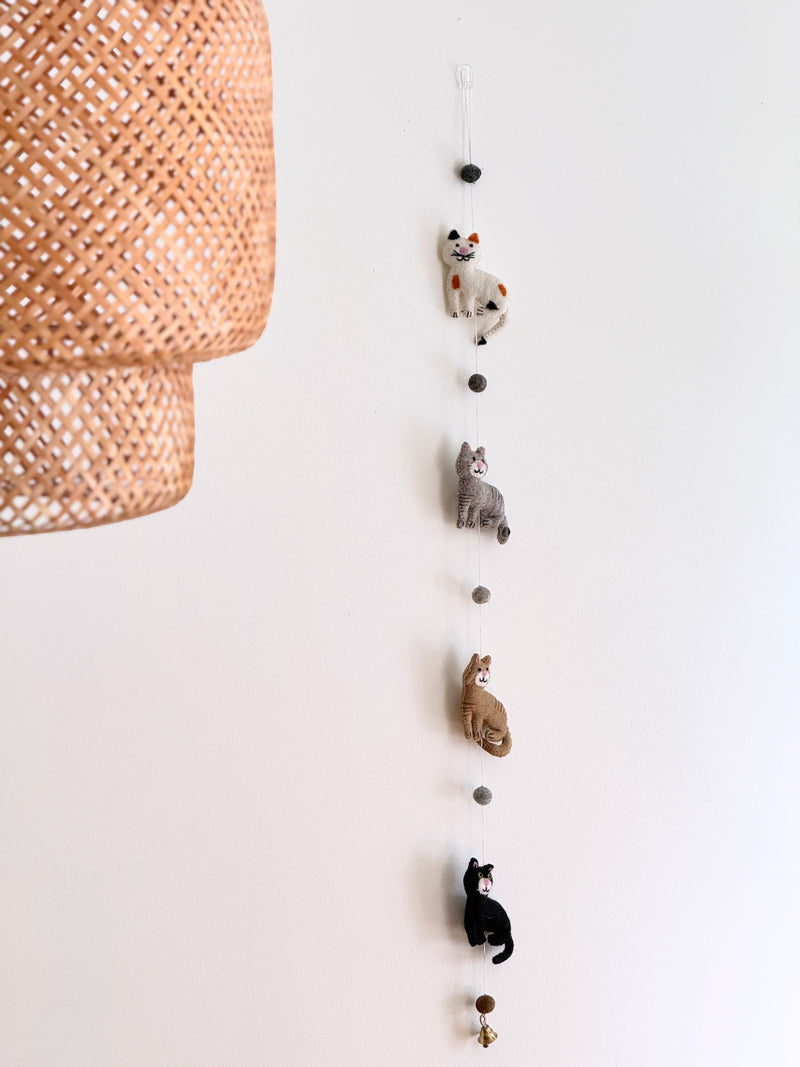 A vibrant wool felt cat garland hangs vertically from a clear plastic hook against a white wall, positioned near a woven lampshade. This 4-foot long garland features four unique 3D felted cats, interspersed with dark gray felt balls. The cats are colored white with black and orange spots, gray, light brown, and black. A small copper bell dangles from the bottom.