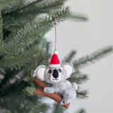 A handcrafted wool felt koala with baby Christmas ornament is displayed hanging on a festive Christmas tree. This charming felt ornament shows a koala wearing a red and white Santa hat, perched on a brown branch with small green leaves. The koala has white inner ears and a black nose, with a jute string attached for hanging.