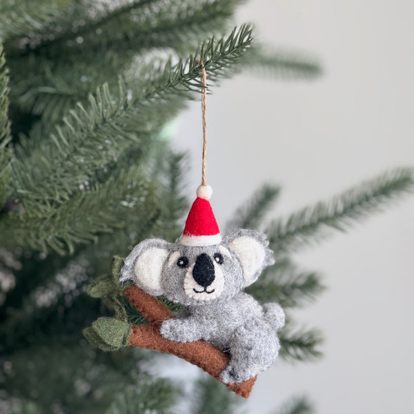 A handcrafted wool felt koala with baby Christmas ornament is displayed hanging on a festive Christmas tree. This charming felt ornament shows a koala wearing a red and white Santa hat, perched on a brown branch with small green leaves. The koala has white inner ears and a black nose, with a jute string attached for hanging.
