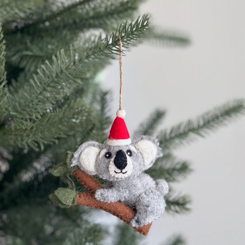 A handcrafted wool felt koala with baby Christmas ornament is displayed hanging on a festive Christmas tree. This charming felt ornament shows a koala wearing a red and white Santa hat, perched on a brown branch with small green leaves. The koala has white inner ears and a black nose, with a jute string attached for hanging.