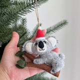 Wool felt koala and baby ornaments are gently presented by a female hand, held alongside a green pine branch. Crafted from gray wool felt, the charming koalas cling to a brown felt branch with green leaves. The main koala wears a red Santa hat with a white pompom. This handcrafted felt Christmas ornament is suspended by a jute string.