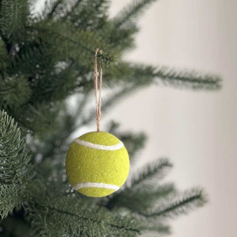A wool felt tennis ball ornament hangs gracefully from a green Christmas tree branch. This handcrafted sports Christmas decoration is bright yellow with a white line encircling its center, accurately mimicking a real tennis ball. A natural jute string is attached, perfect for adding a playful touch to your holiday decor.