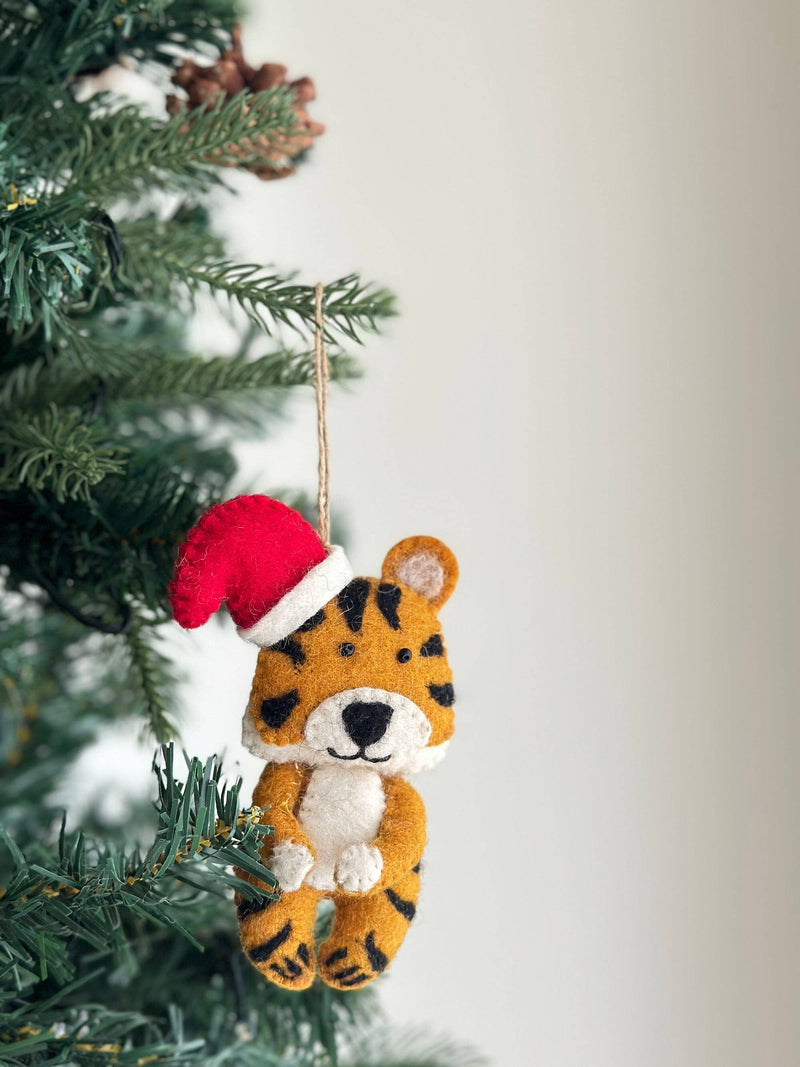 A felt tiger ornament with a red Christmas hat is displayed on a green Christmas tree. This handcrafted felt tiger ornament features a tan body with black stripes and is suspended by a jute string, enhancing its appeal as a unique Christmas tree ornament amidst the festive setting.