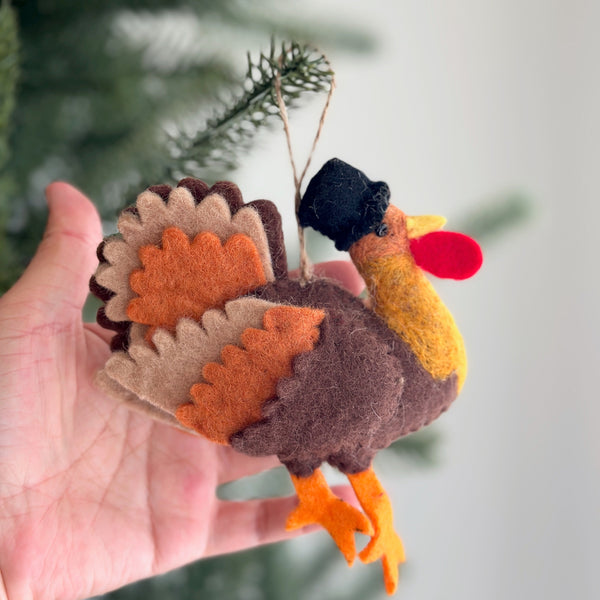 A wool felt turkey ornament is held in a female hand, set against a blurred green Christmas tree. The plump brown turkey ornament features a black hat, layered tail feathers in brown, tan, and orange, an orange beak, and orange feet. A jute string is attached for hanging, evoking a cozy, festive holiday mood.