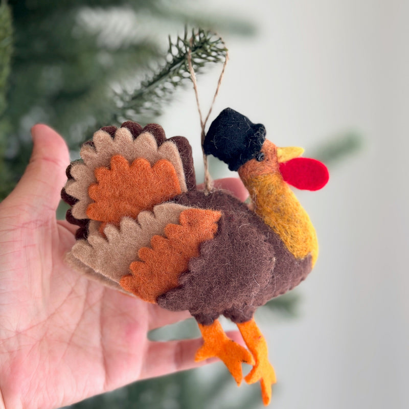 A wool felt turkey ornament is held in a female hand, set against a blurred green Christmas tree. The plump brown turkey ornament features a black hat, layered tail feathers in brown, tan, and orange, an orange beak, and orange feet. A jute string is attached for hanging, evoking a cozy, festive holiday mood.